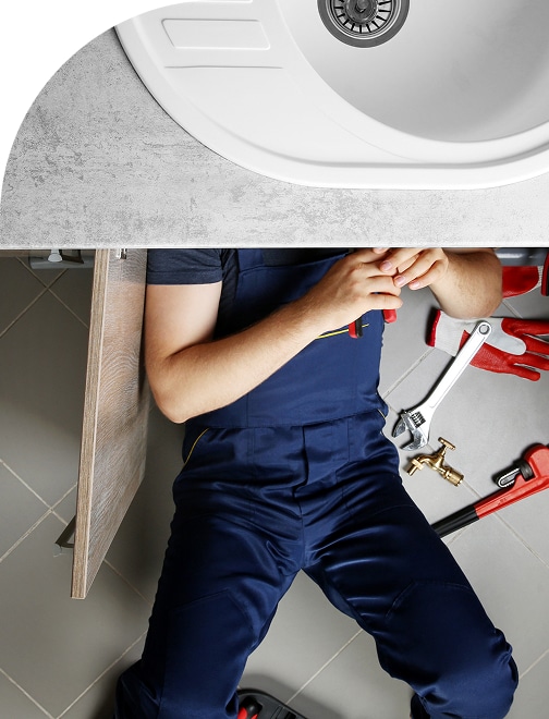 Plombier en salopette bleue travaillant sous un évier blanc neuf, entouré de clés et d'un robinet en laiton sur carrelage gris.