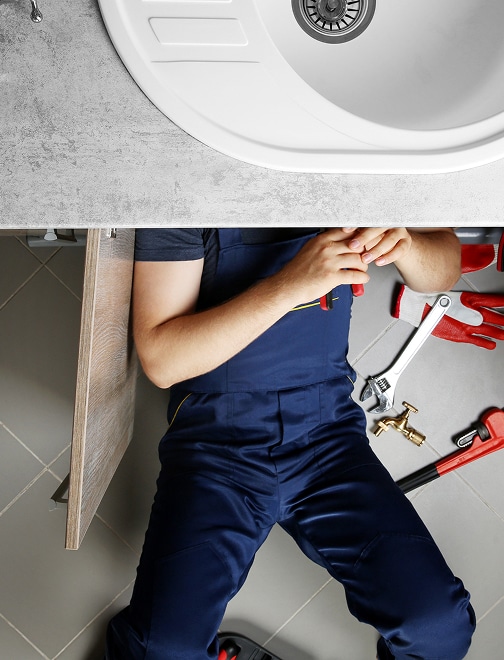 Réparation évier : plombier sous le lavabo avec outils Plombier en combinaison bleue réparant sous un évier de cuisine blanc, outils comme clé à molette et robinet à côté de lui.