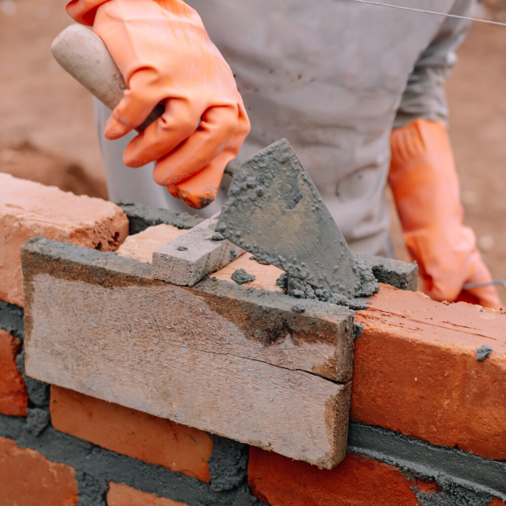 Maçonnerie : Construction de mur en briques avec truelle Maçon portant des gants orange utilisant une truelle pour appliquer du mortier sur une nouvelle rangée de briques rouges, avec un fil de maçonnerie visible.