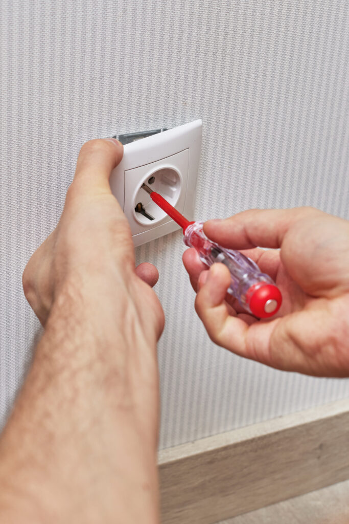 Test de tension prise électrique avec tournevis testeur Un électricien vérifie une prise murale blanche européenne avec un tournevis testeur à manche rouge et transparent, contre un mur texturé clair.