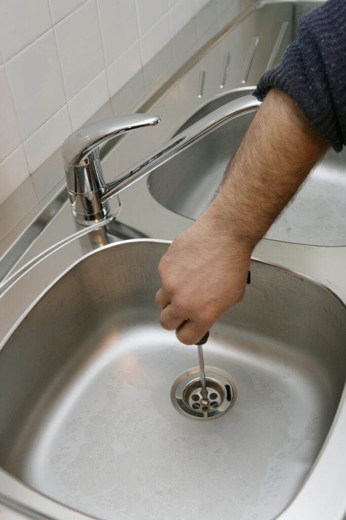 Réparation évier : homme débouchant le siphon avec un tournevis Main avec un pull bleu foncé insérant un tournevis dans le siphon d'un évier de cuisine en acier inoxydable.