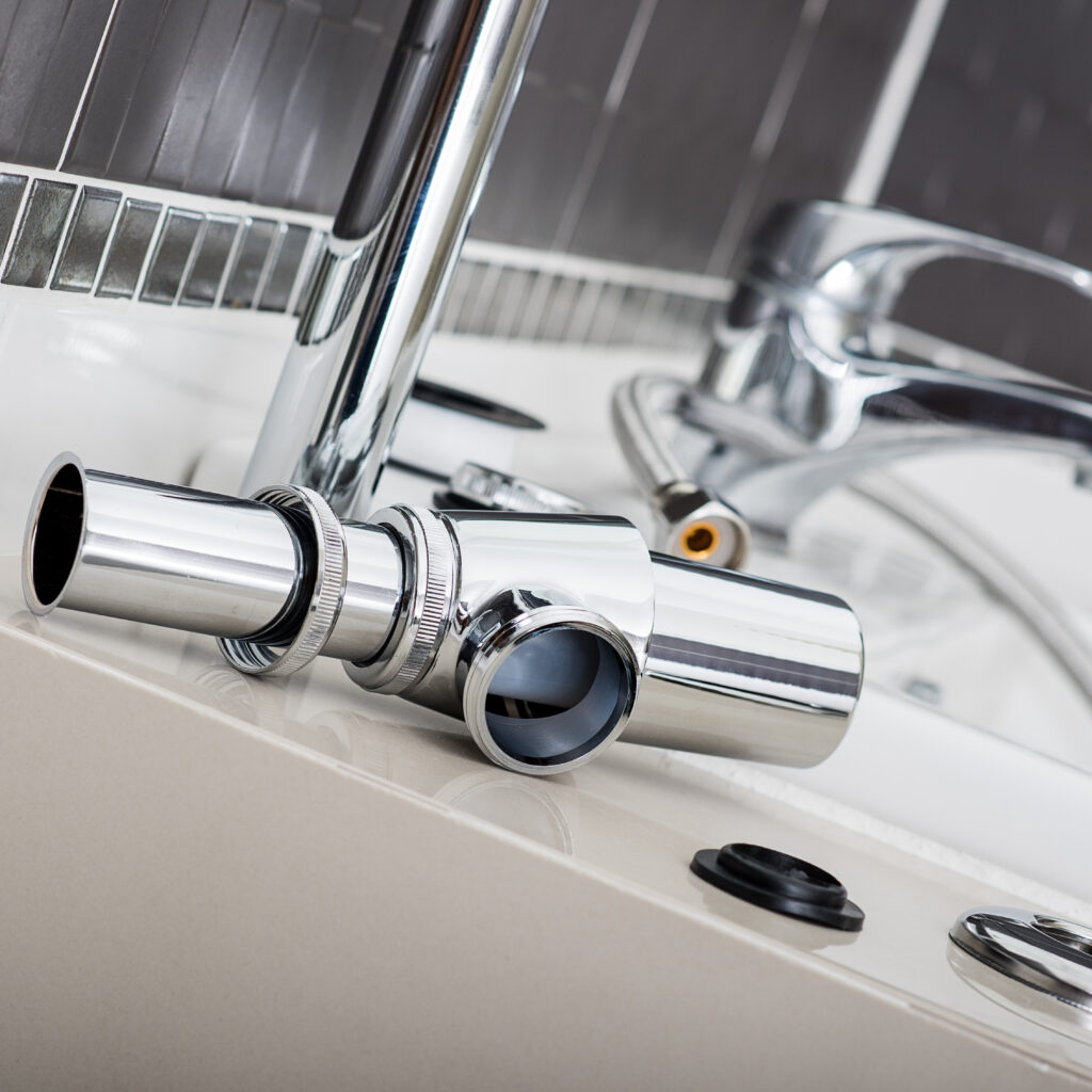 Installation Siphon Chromé Lavabo : Pièces Détachées Assemblage d'un siphon chromé neuf pour lavabo, posé sur une vasque claire, avec robinetterie et carreaux muraux gris en arrière-plan.