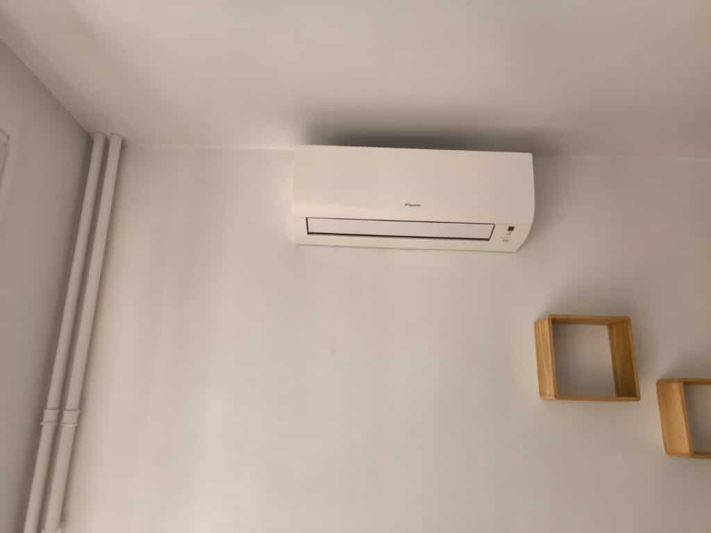 Climatiseur mural blanc Daikin installé sur un mur blanc, près de tuyaux verticaux et de deux petites étagères cubiques en bois clair.