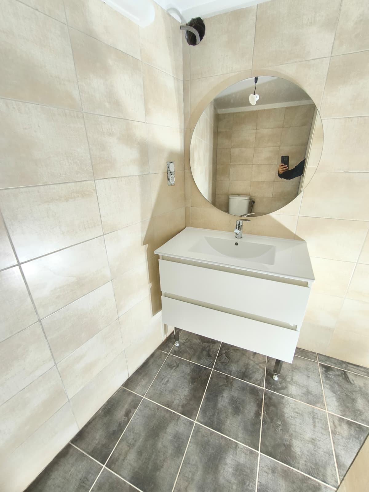 Salle Bain Moderne: Meuble Lavabo, Miroir Rond et Carrelage Salle de bain moderne avec meuble-lavabo blanc suspendu, miroir rond, carrelage mural beige clair et sol gris foncé. Prise électrique visible.